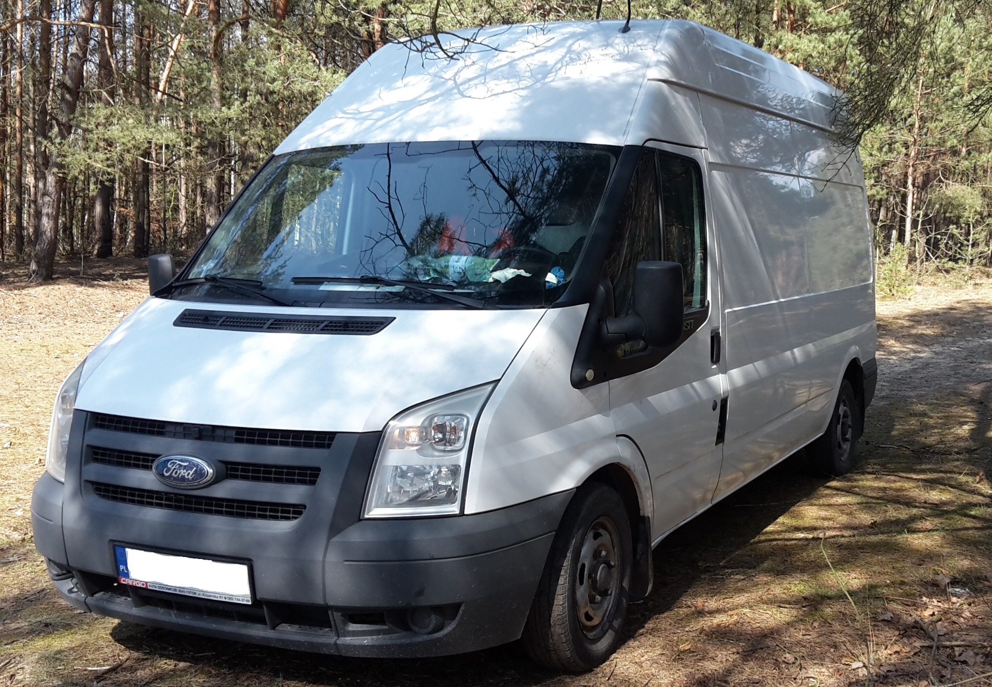 Biały samochód dostawczy Ford Transit na leśnej polanie, widok z przodu, na przedniej szybie odbijają się korony drzew.