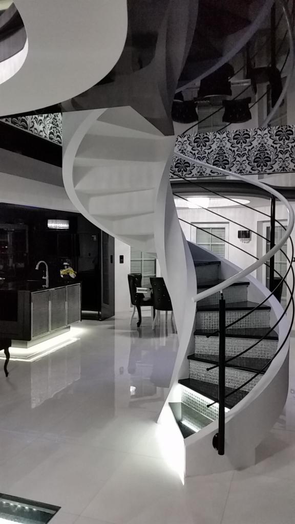 Białe schody spiralne z czarnymi stopniami i metalową balustradą w nowoczesnym wnętrzu z błyszczącą podłogą i widoczną kuchnią w tle.