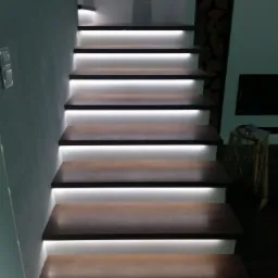 Nowoczesne schody z ciemnego drewna z podświetleniem LED, widok z dołu, w tle ściana z ułożonym drewnem kominkowym.