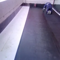 Pracownik na kolanach, wygładza papę na płaskim dachu balkonu przed zabudową, widoczny laserowy niwelator, częściowo ułożona izolacja termiczna.