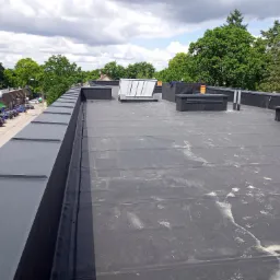 Czarny, płaski dach pokryty papą termozgrzewalną z widocznymi śladami użytkowania, kominami wentylacyjnymi i świetlikiem dachowym, na tle zabudowy miejskiej i zieleni.