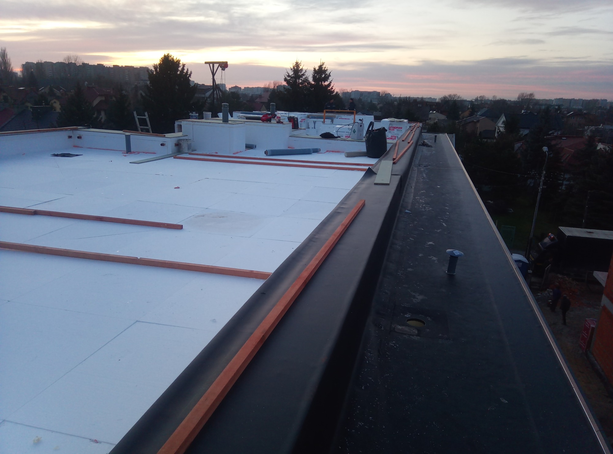 Płaski dach w trakcie izolacji styropianem, widoczne drewniane legary i czarna papa, w tle zabudowa miejska o zmierzchu.