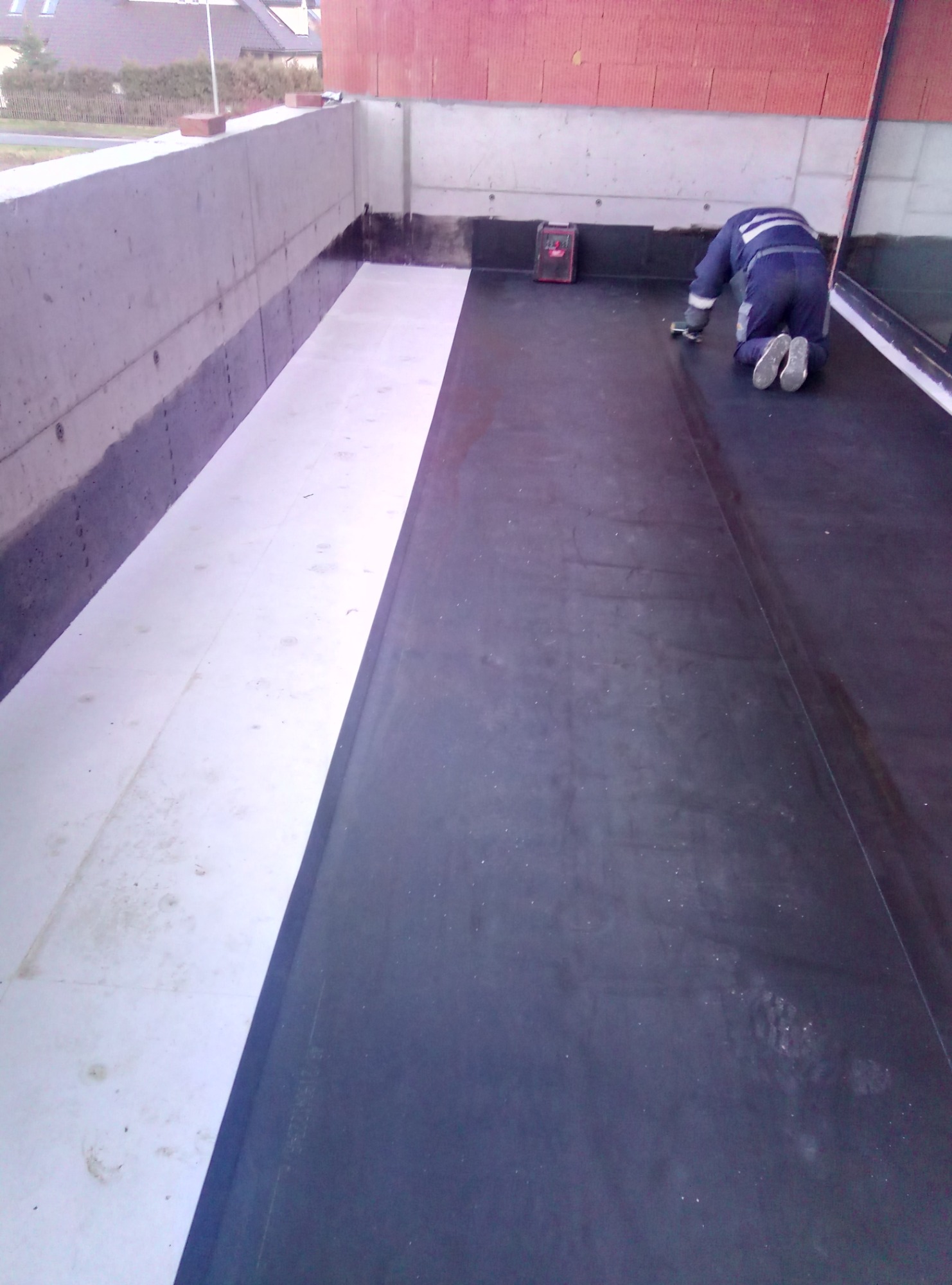 Pracownik na kolanach, wygładza papę na płaskim dachu balkonu przed zabudową, widoczny laserowy niwelator, częściowo ułożona izolacja termiczna.