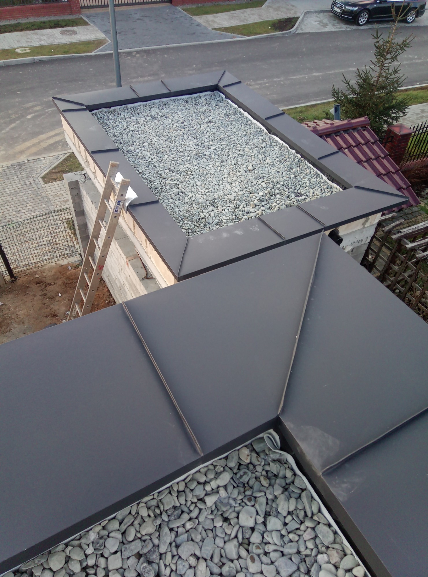 Widok z góry na płaski dach z czarnej blachy z fragmentami wypełnionymi szarym żwirem, drabina oparta o ścianę budynku w tle.