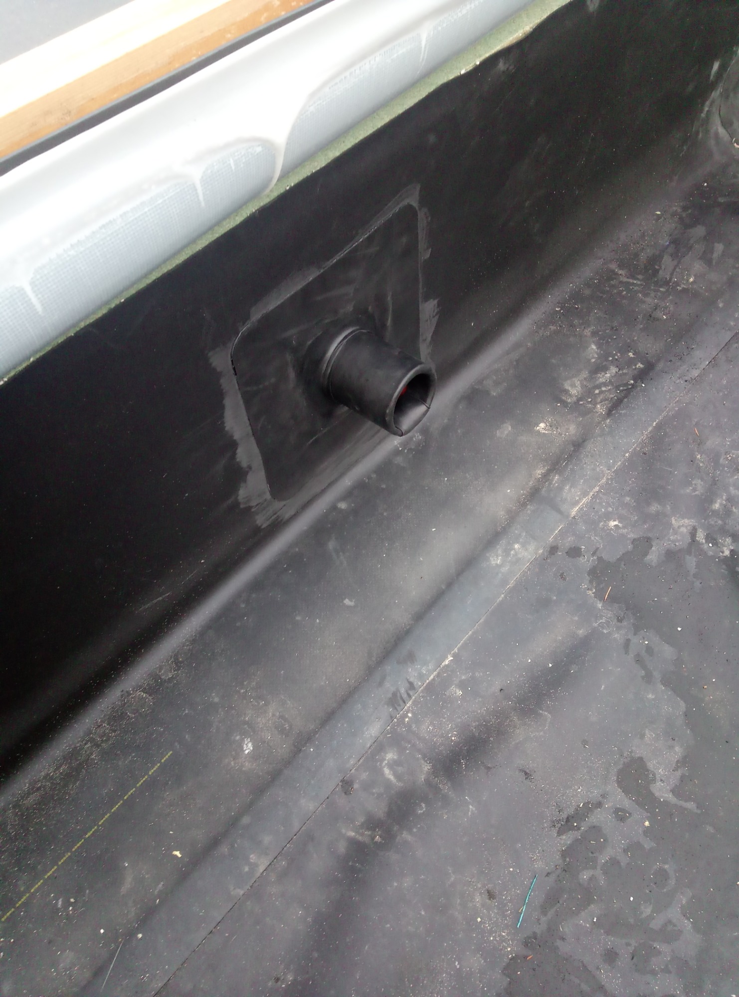 Czarny, gumowy odpływ zamontowany na płaskiej powierzchni dachu z papy bitumicznej, widoczne ślady uszczelniania wokół rury.