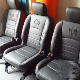 Trzy czarne, skórzane fotele samochodowe z wyhaftowanym logo marki na oparciach, ustawione w warsztacie przed ścianą.