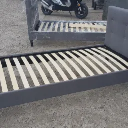 Szara tapicerowana rama łóżka z drewnianym stelażem, ustawiona na betonowym podłożu, z drugim podobnym łóżkiem w tle, częściowo owiniętym folią, oraz skuterem w tle.