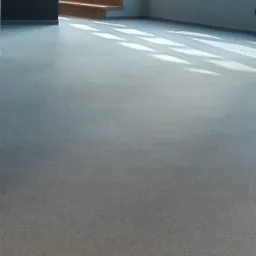 Jasnoszara, jednolita powierzchnia podłogi w pustym pomieszczeniu biurowym z dużymi oknami i drewnianymi schodami w tle.