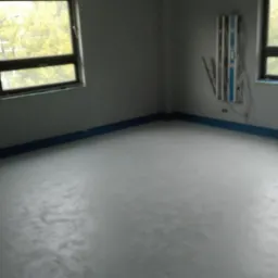 Nowa, szara posadzka jastrychowa w jasnym, pustym pokoju z dwoma oknami, zabezpieczona niebieską folią przy ścianach.