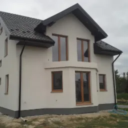 Elewacja domu jednorodzinnego po ociepleniu, z widocznymi oknami w drewnianych ramach i ciemną dach&oacute;wką. Na ziemi leży zwinięty wąż ogrodowy.