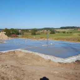 Świeżo wylana, lśniąca posadzka fundamentowa na działce budowlanej, widok na wiejski krajobraz z pagórkami i lasem w tle.