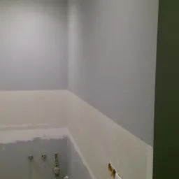 Wnętrze łazienki w trakcie remontu: szare ściany częściowo pokryte białym gruntem, widoczne instalacje wodne i elektryczne, surowe wykończenie.