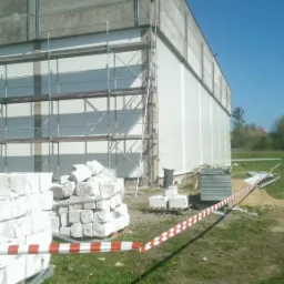 Budynek przemysłowy w trakcie ocieplania elewacji, widoczne rusztowania, materiały izolacyjne i taśma ostrzegawcza na trawniku.