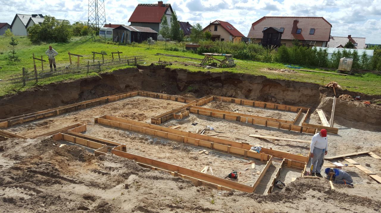 Przygotowanie fundamentów pod budowę domu: wykop z drewnianym szalunkiem, widoczni dwaj pracownicy, jeden w czerwonej czapce, drugi w niebieskim kombinezonie, w tle zabudowa jednorodzinna.