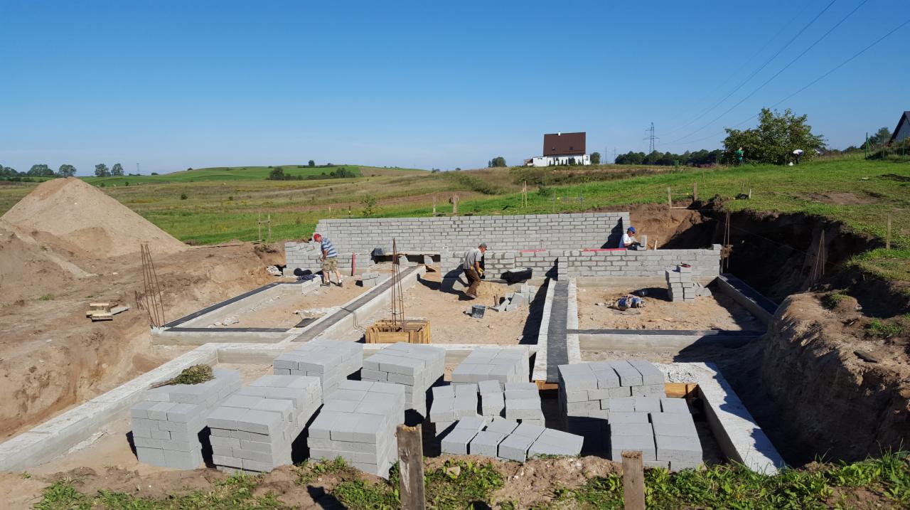 Budowa fundamentów domu z bloczków betonowych na wsi, widoczni pracownicy i stos materiałów budowlanych.