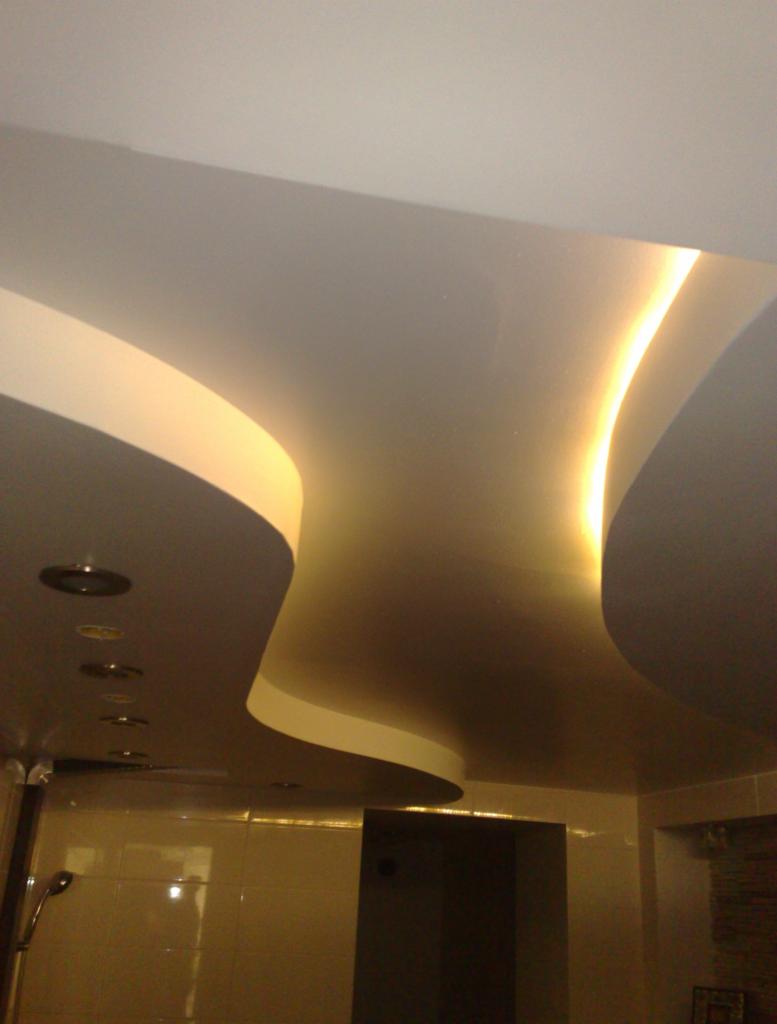 Błyszczący, podwieszany sufit w łazience z oświetleniem LED, tworzący falisty wzór. Widoczne halogeny i fragment kabiny prysznicowej.