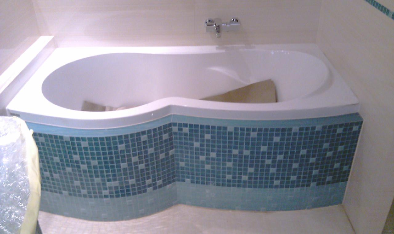 Wanna obudowana mozaiką w odcieniach błękitu i bieli. Widoczna chromowana bateria. Jasne płytki na ścianach i podłodze. Nietypowy kształt wanny.