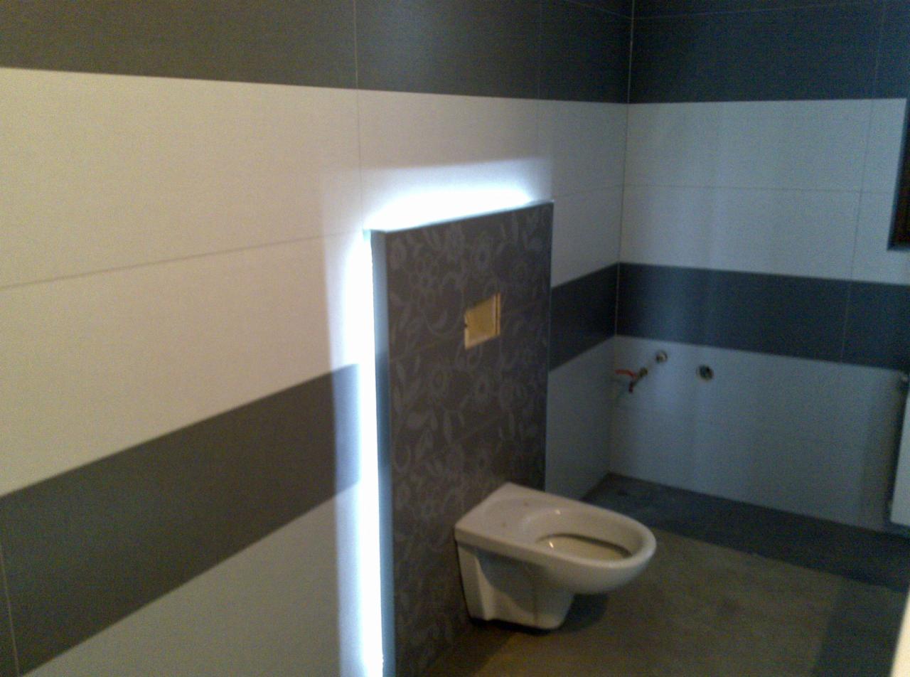 Świeżo wyremontowana łazienka z częściowo zamontowaną toaletą wiszącą i wykończonymi płytkami w odcieniach szarości i bieli w nowoczesnym stylu.