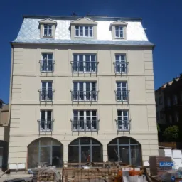 Wielopiętrowy budynek z beżową elewacją, srebrnym dachem i balkonami, w trakcie prac wykończeniowych, widoczne materiały budowlane na pierwszym planie.