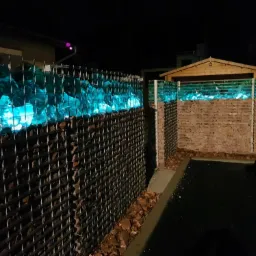 Gabionowa ściana z podświetlonymi na niebiesko szklanymi elementami, widoczna w nocy na tle budynku i drewnianego domku.
