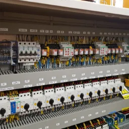 Rozbudowana szafa sterownicza z licznymi zabezpieczeniami i przekaźnikami, oznaczona etykietami, z widocznym okablowaniem i tabliczką ostrzegawczą o wysokim napięciu.