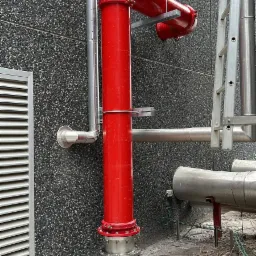 Czerwona rura hydrantowa z zaworami i mocowaniami przy ścianie budynku, obok drabina i inne instalacje.