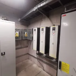 Instalacja elektryczna w pomieszczeniu technicznym: szafa sterownicza ABB, korytka kablowe pod sufitem, zabezpieczenia elektryczne w metalowej klatce.