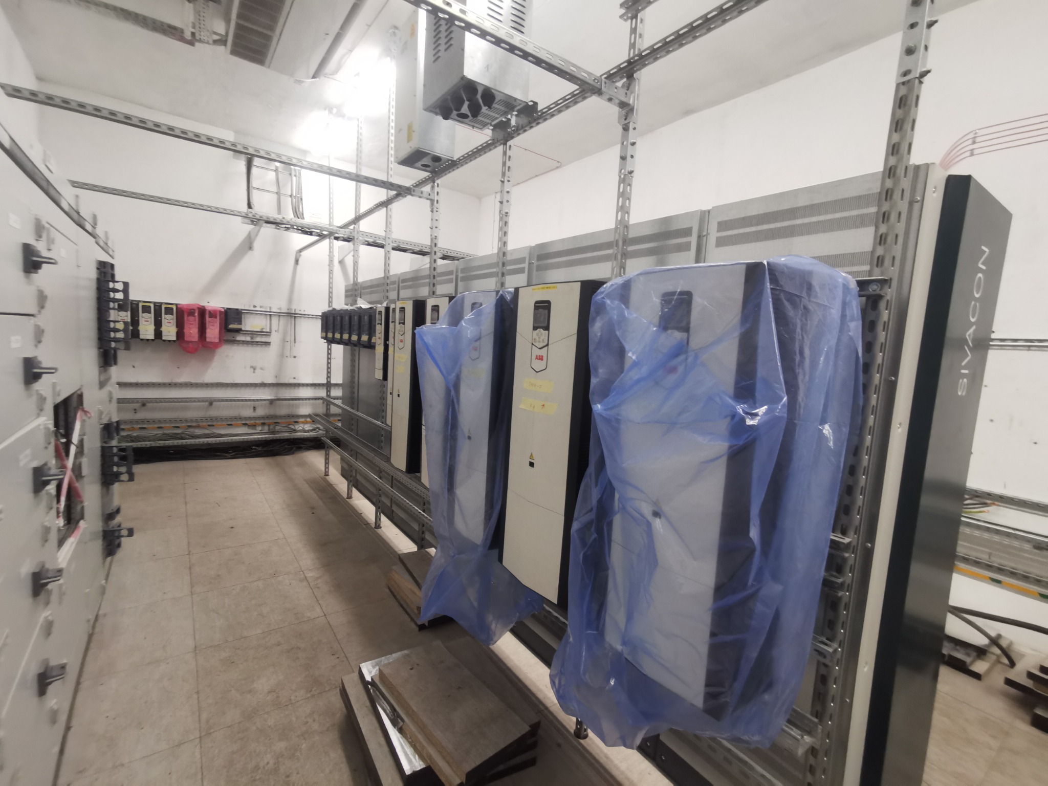 Wnętrze hali przemysłowej z zainstalowanymi, częściowo osłoniętymi folią, szafami sterowniczymi ABB, zamontowanymi na metalowych konstrukcjach nośnych.