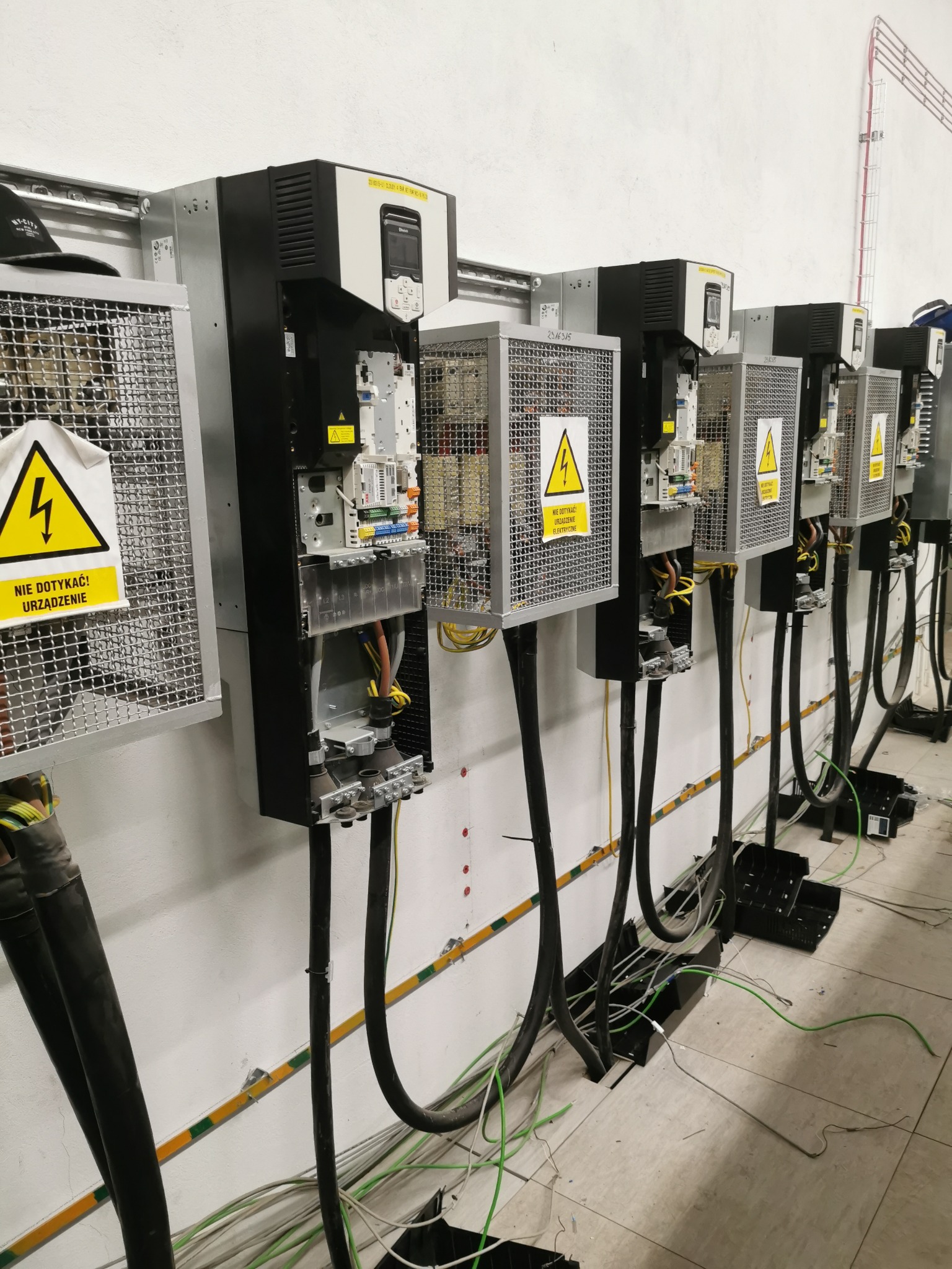 Rząd zainstalowanych, otwartych szaf sterowniczych z ostrzeżeniami o napięciu, podłączonych grubymi kablami, widoczne kable na podłodze.
