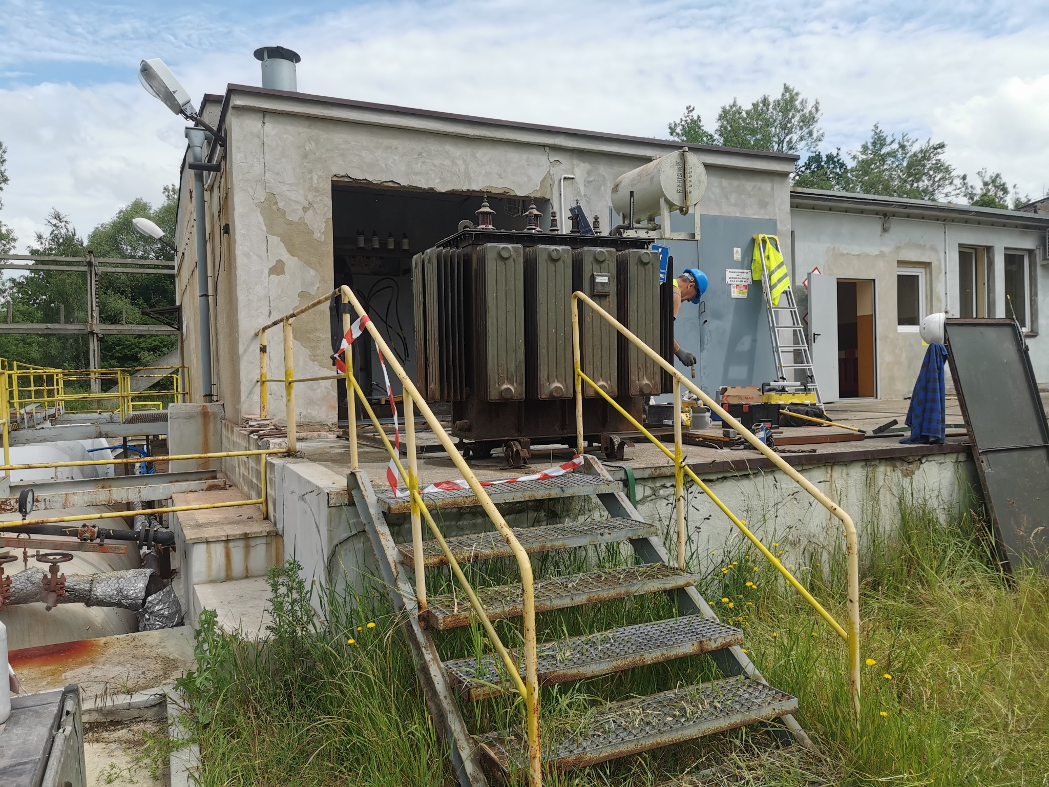 Pracownik w niebieskim kasku przy transformatorze na zewnątrz budynku przemysłowego, widoczne schody z żółtą barierką i taśma ostrzegawcza.