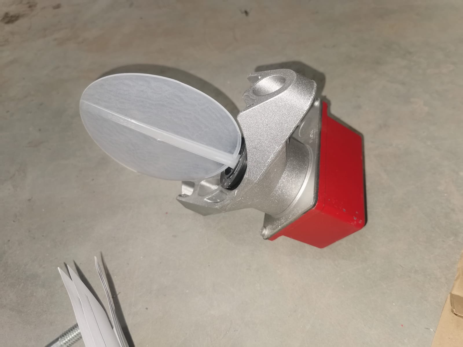 Szlifowany, srebrny element maszyny z czerwonym akcentem, zabezpieczony plastikową osłoną, leżący na szarej powierzchni.