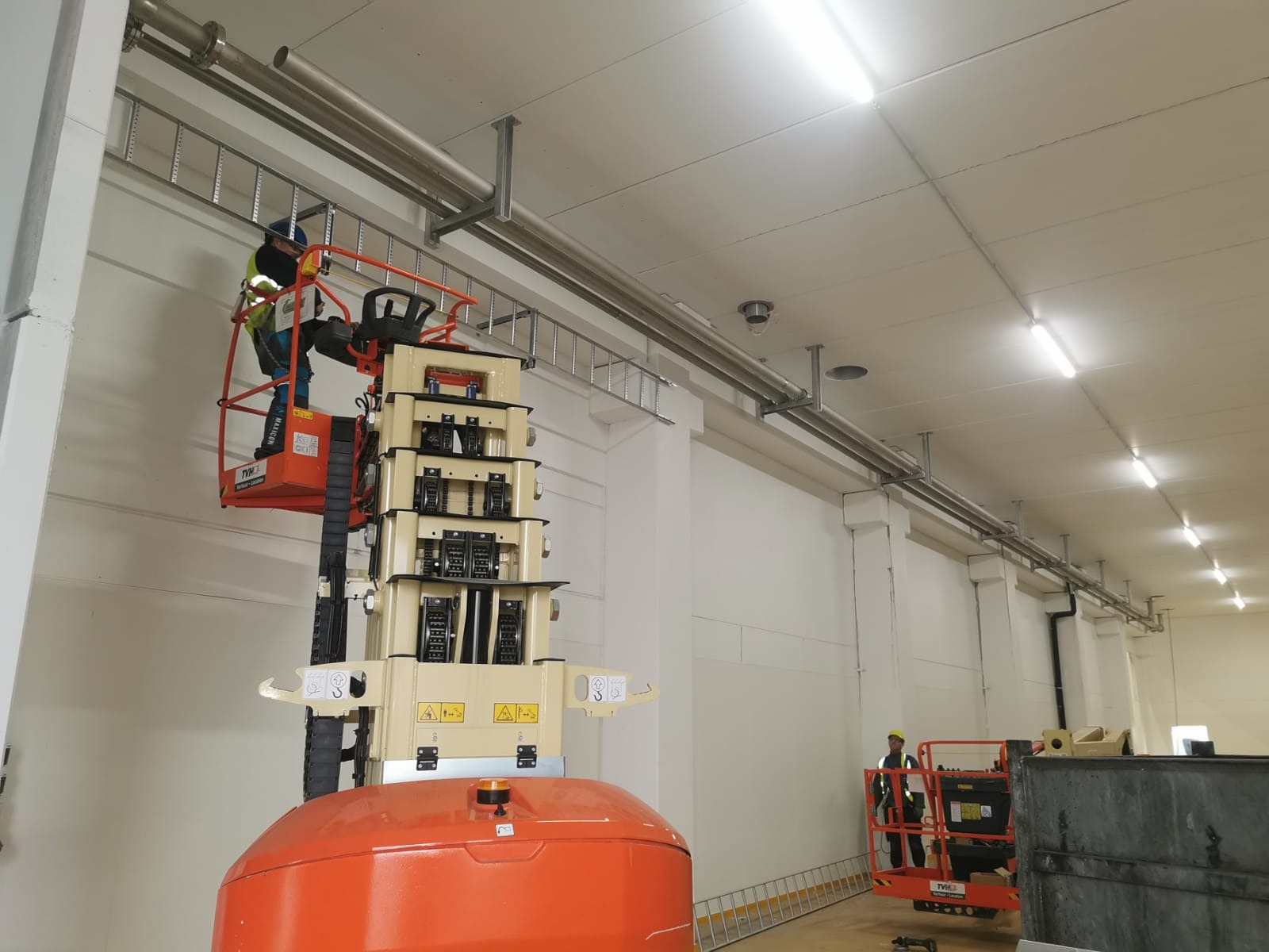 Montaż instalacji pod sufitem w hali produkcyjnej z użyciem podnośnika koszowego; widoczne rury, koryta kablowe i oświetlenie.