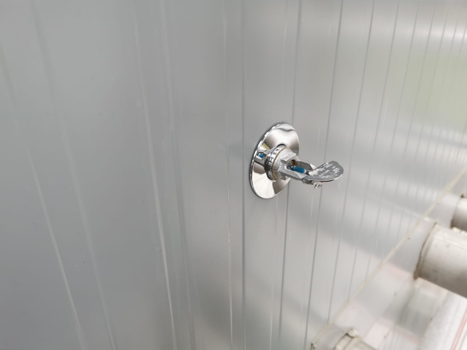 Chromowany zraszacz przeciwpożarowy zamontowany na szarej, panelowej ścianie w hali, widoczne rury instalacji w tle.