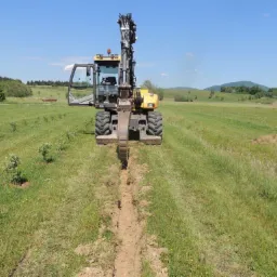 Żółta koparka kołowa tworzy wąski wykop w zielonym terenie, widok z przodu, błękitne niebo. Nowe nasadzenia drzew w tle. Drenaż na łące.