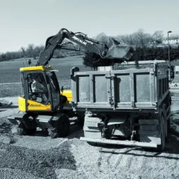 Żółta koparka Mecalac ładuje gruz do ciężarówki na placu budowy. W tle widoczne budynki mieszkalne i drzewa. Częściowo odbarwione zdjęcie z żółtymi akcentami.