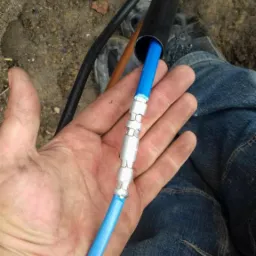 Dłoń trzyma niebieski przewód z metalowymi złączkami, prowadzący do czarnej rury w ziemi, widoczne buty i dżinsy osoby wykonującej pracę.