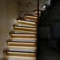 Drewniane schody z podświetleniem LED na stopniach, ściana z cegły w tle, nowoczesny design.