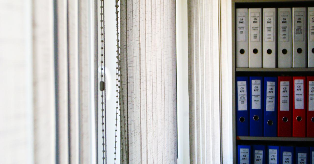 Półka z segregatorami w biurze, widoczne rolety pionowe. Segregatory w kolorach białym, niebieskim i czerwonym, z nadrukami. Neutralne, stonowane tło.