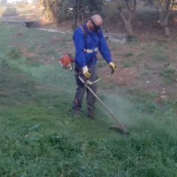 Pracownik w niebieskim kombinezonie i okularach ochronnych kosi trawę przy użyciu spalinowej podkaszarki na tle drzew i betonowego przepustu.