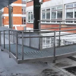 Zewnętrzna platforma z metalową kratą i stalowymi barierkami, zapewniająca dostęp dla osób z niepełnosprawnościami, zlokalizowana przed budynkiem z oknami i tabliczką informacyjną.