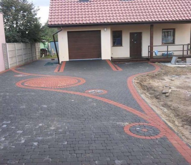 Ułożona kostka brukowa w kolorze szarym z czerwonymi akcentami tworzącymi wzory przed domem z garażem i tarasem. Widok z góry na gotową realizację.