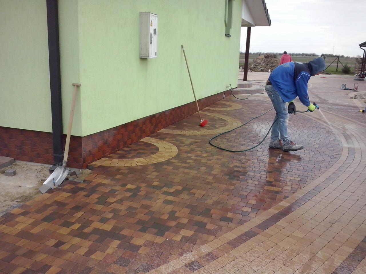 Układanie mozaiki brukowej w kształcie fali. Pracownik czyści powierzchnię wodą pod ciśnieniem. Widoczna łopata i szczotka przy ścianie budynku.