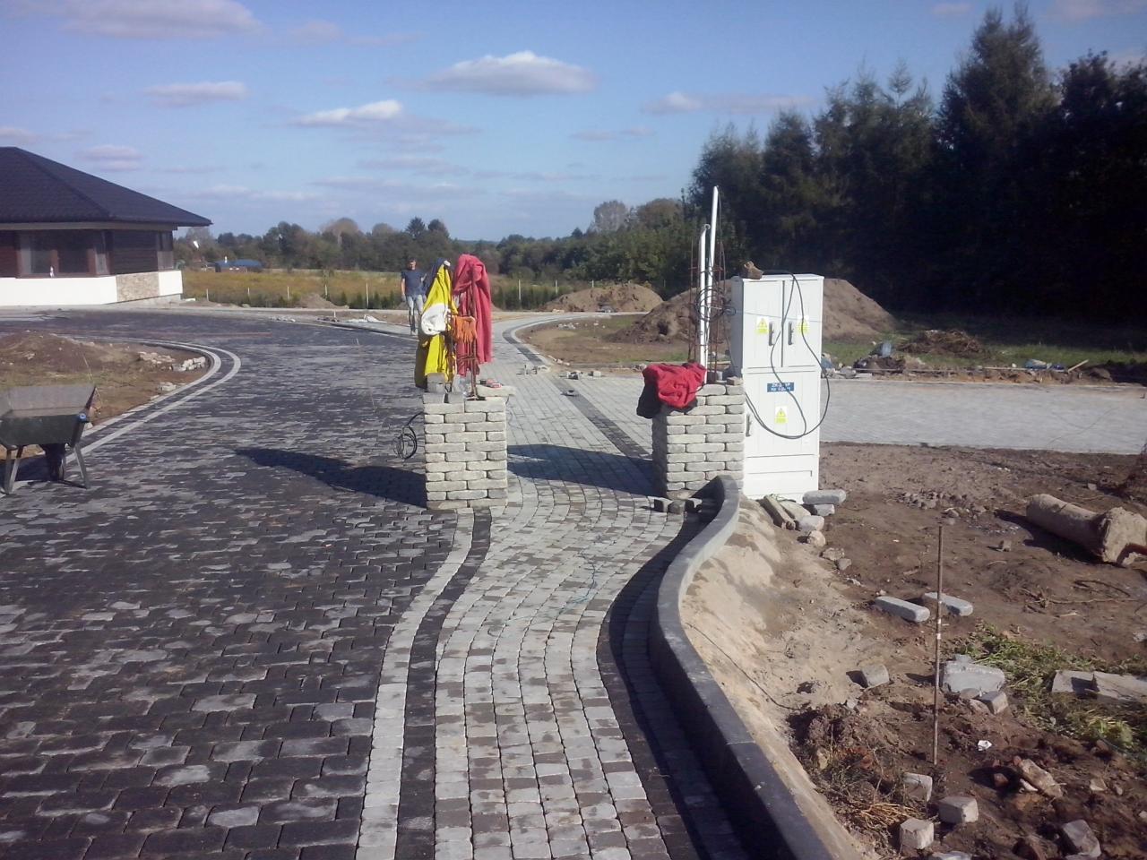 Ułożona, dwukolorowa kostka brukowa na posesji z budynkiem w tle. Widoczne słupki z odzieżą roboczą i skrzynka elektryczna. Częściowo ułożona kostka po prawej stronie.