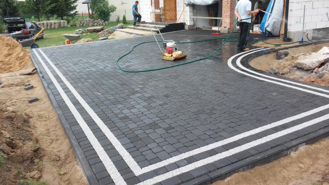 Ułożona, ciemnoszara kostka brukowa z dwoma białymi pasami. W tle widoczna ubijarka, dom w budowie i pracownicy. Całość sprawia wrażenie świeżo ułożonej nawierzchni.