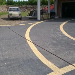 Ułożona, szara kostka brukowa z żółtymi, łukowatymi pasami. W tle budynek z kolumnami, samochody i kamienny murek.