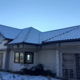 Niedokończony dom jednorodzinny z szarą dachówką ceramiczną pokrytą cienką warstwą śniegu, widoczne okna z folią zabezpieczającą, ściany z białego bloczka, drewniane palety przy ścianie bocznej.