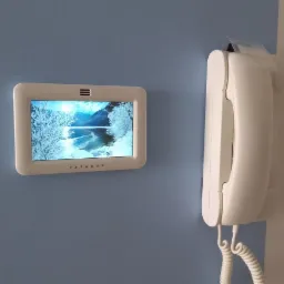 Biały panel dotykowy z wyświetlaczem przedstawiającym zimowy krajobraz, zamontowany na niebieskiej ścianie obok białego domofonu z kręconym kablem.