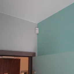 Biały czujnik ruchu zamontowany w rogu pokoju, nad futryną drzwi, na styku szarej i turkusowej ściany.