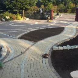 Ukończony podjazd i ścieżka z kostki granitowej w ogrodzie, z dekoracyjnymi wzorami i świeżo przygotowanymi rabatami ziemnymi. Częściowo widoczna osoba.