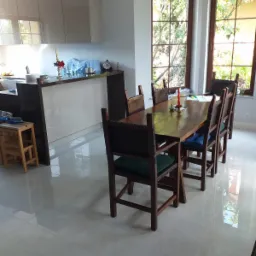 Jasna kuchnia z beżowymi szafkami i drewnianym stołem z krzesłami na błyszczącej, jasnej podłodze. Widoczne dwa okna z widokiem na zieleń na zewnątrz.